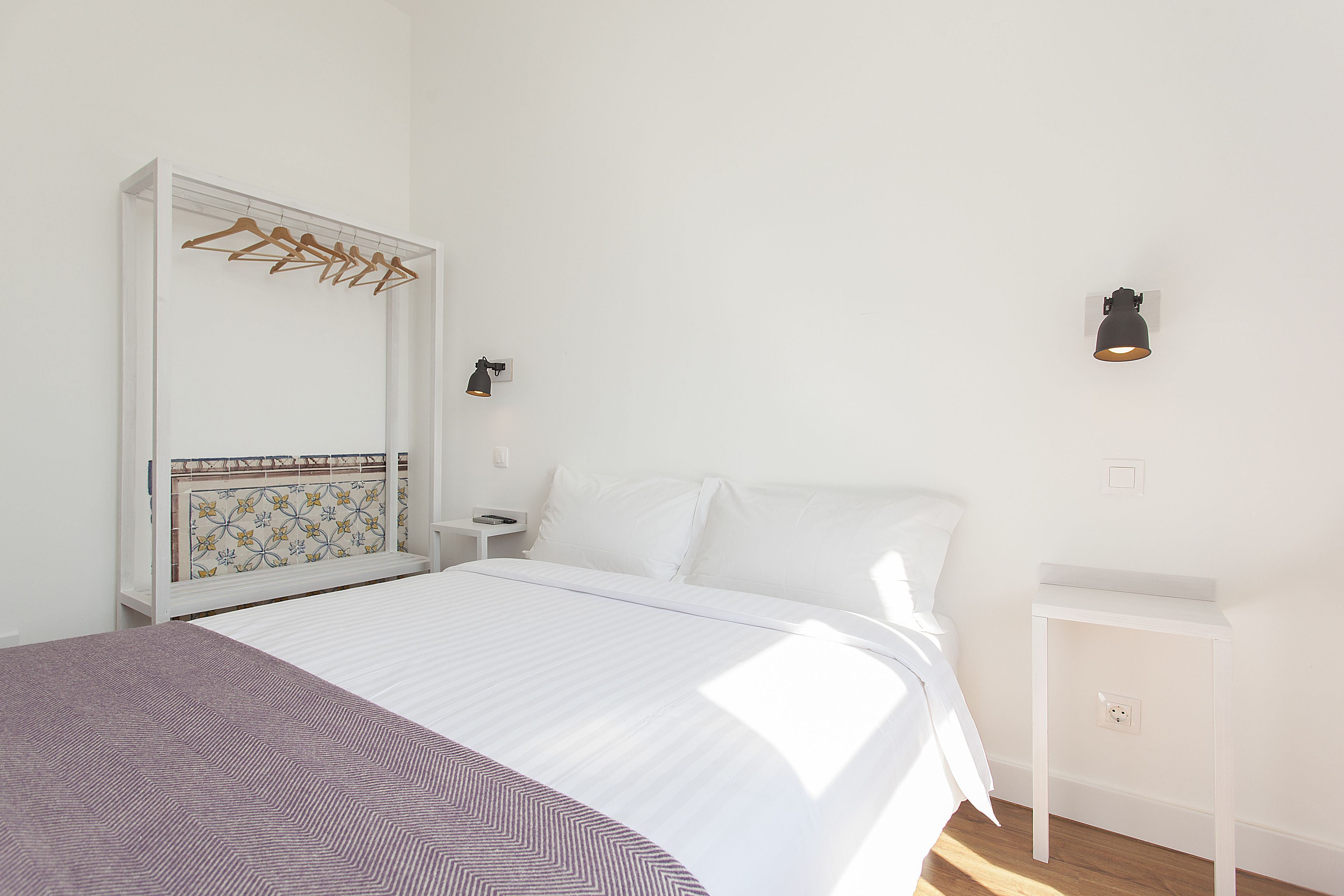 A bedroom with a bed, a bedside table, a wall-mounted lamp, and a coat rack.