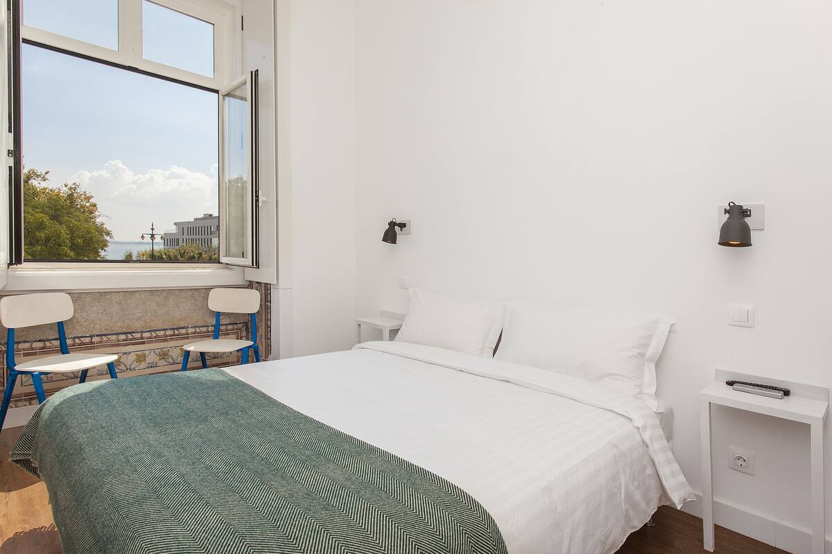 A hotel room with a large window, a bed with white linens, a green patterned throw, two white nightstands, and a view of the cityscape.