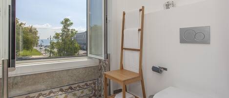 A bathroom with a toilet, a wooden drying rack, a window with a view of the outside, and a tiled floor.