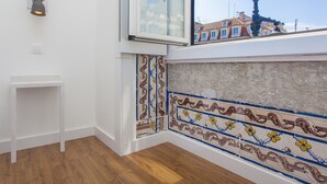 Een hoek van een kamer met een witte muur, een klein wit tafeltje, een decoratieve tegelrand en een raam met uitzicht op een gebouw.