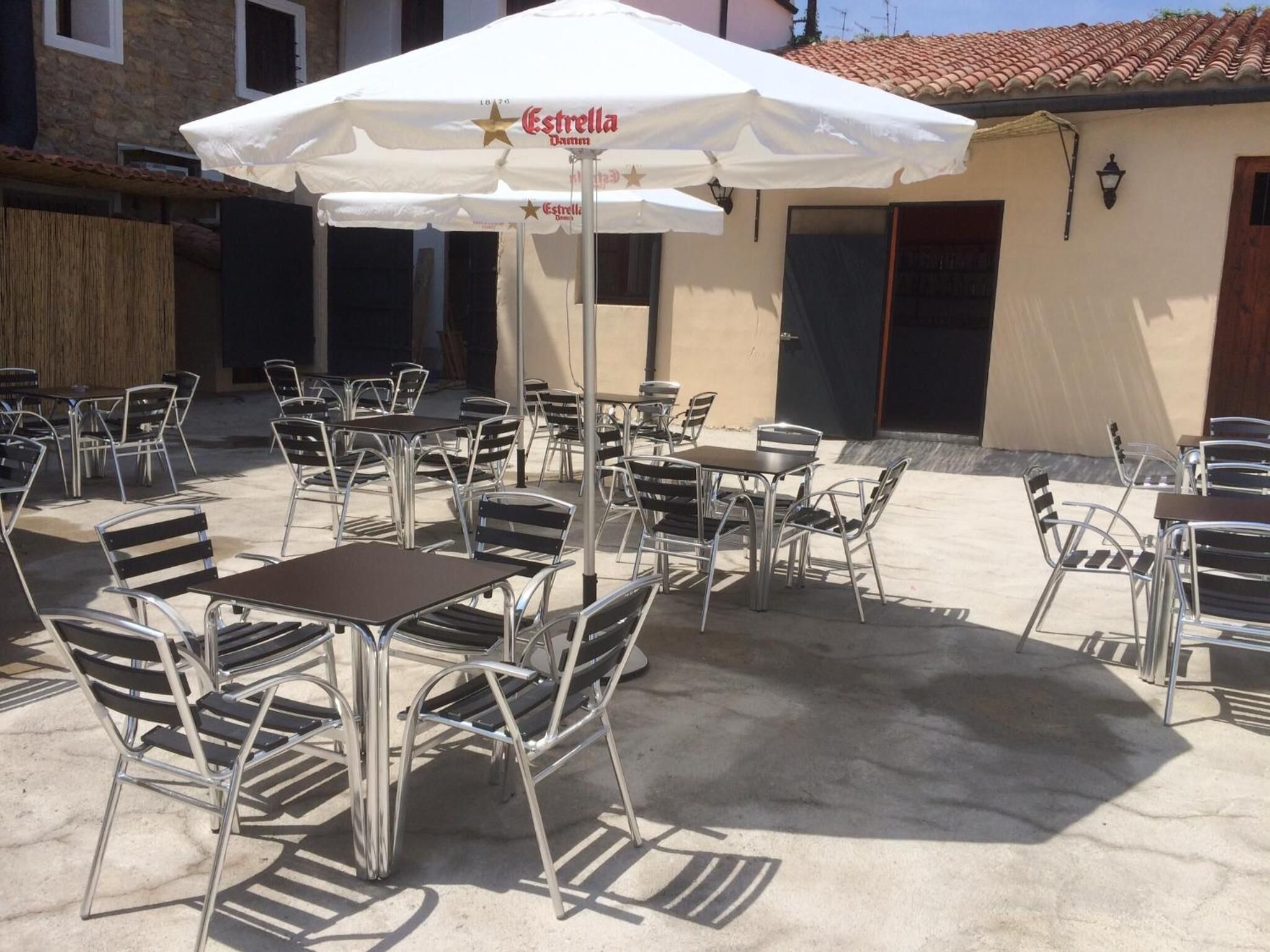 An outdoor patio area with metal tables and chairs, a white umbrella, and a building with a tiled roof in the background.