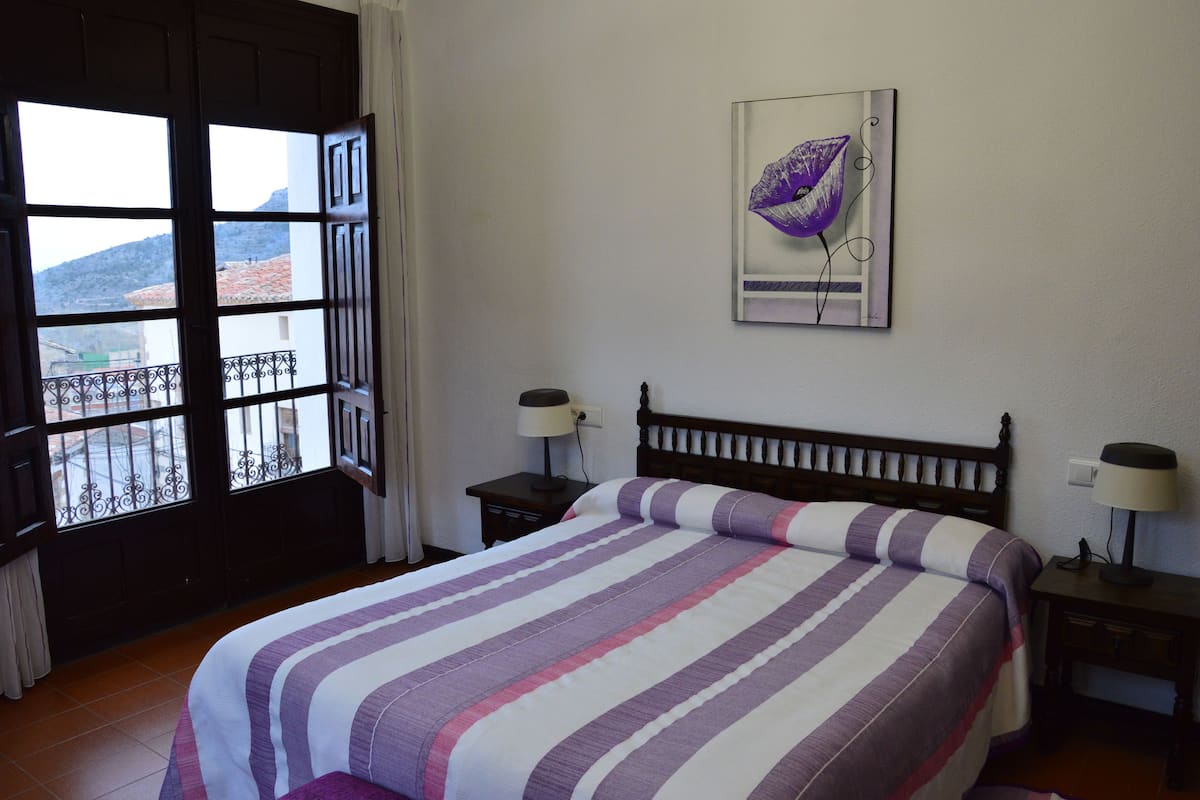 Een slaapkamer met een bed, twee nachtkastjes met lampen, een raam met uitzicht op gebouwen en bergen, en een kunstwerk aan de muur.