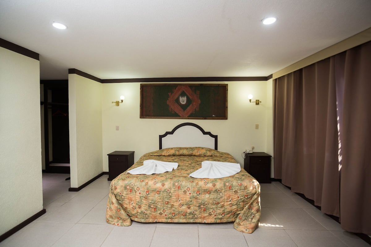 A bedroom with a bed, two bedside tables, a nightstand, a painting on the wall, and a curtain on the window.