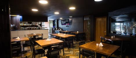 A restaurant interior with multiple wooden tables and chairs, a bar area, and a television screen on the wall.