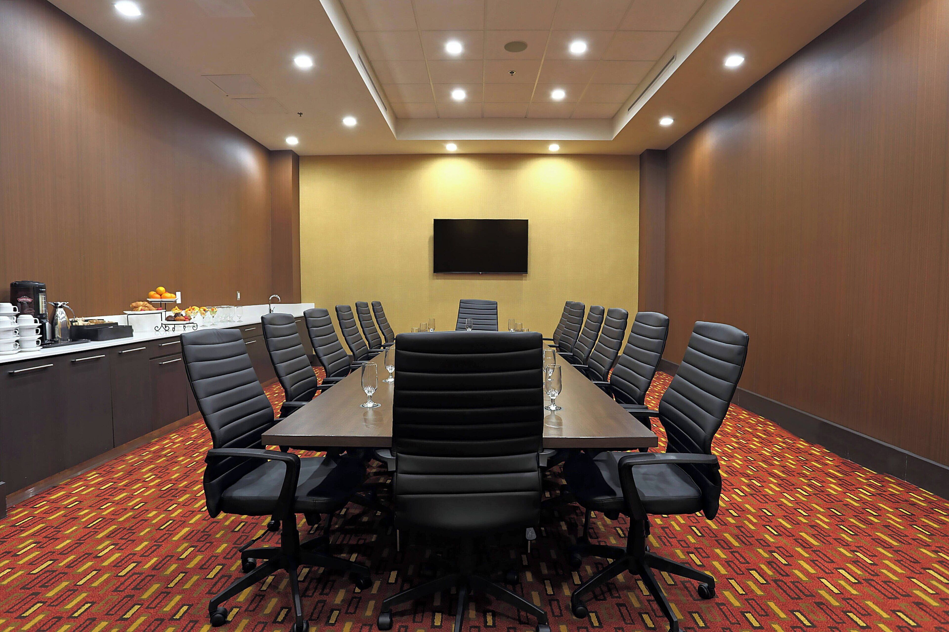 A conference room with a long table, black chairs, a flat-screen TV, and a coffee station.