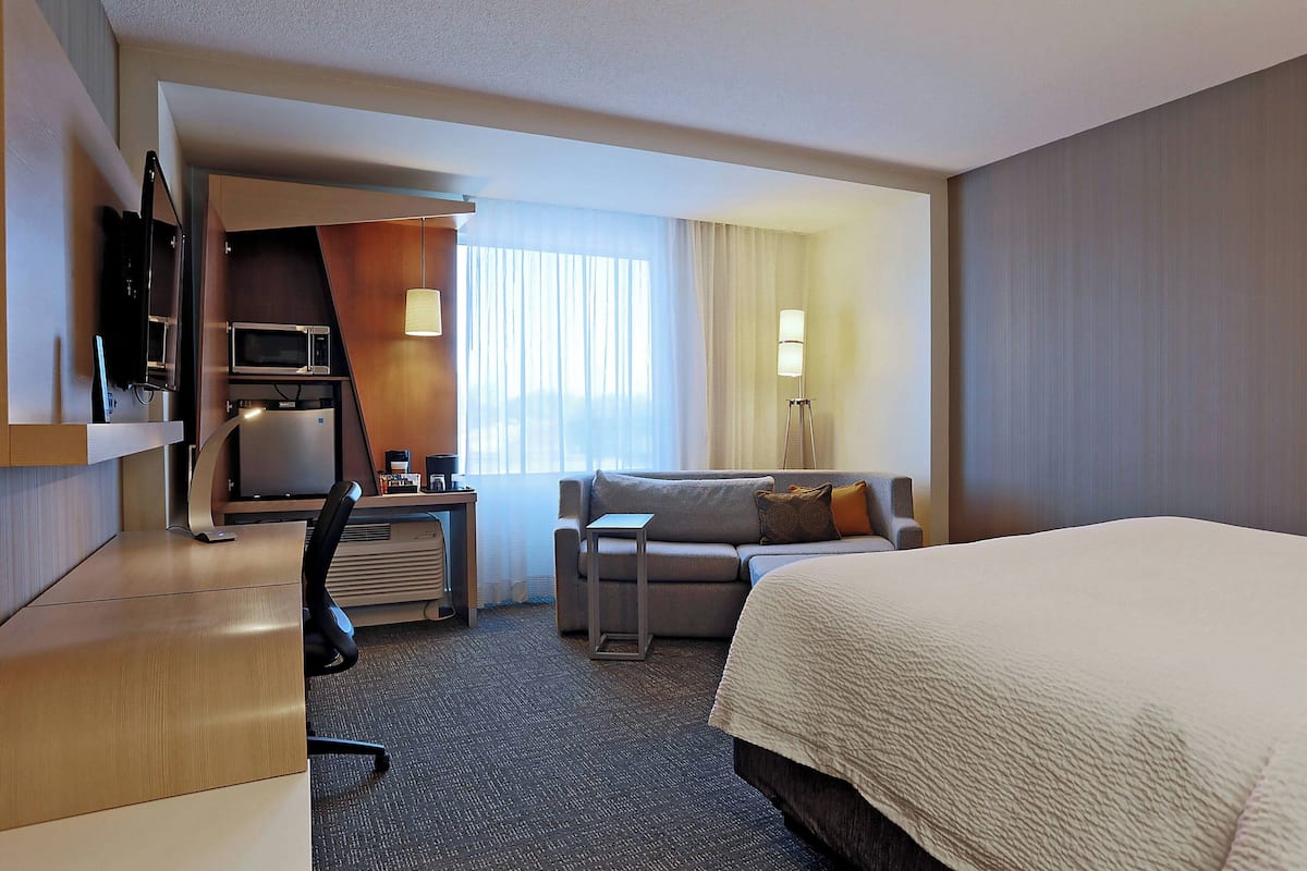 A hotel room with a bed, a desk with a computer, a microwave, a TV, a sofa, and a lamp.