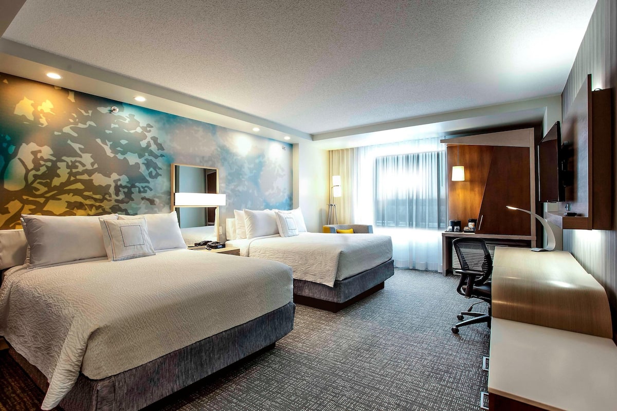 A hotel room with two beds, a desk, a chair, a TV, and a large window with curtains.