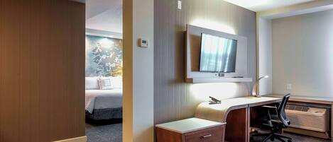 A hotel room with a bed, a desk with a chair, a television mounted on the wall, and a thermostat on the wall.