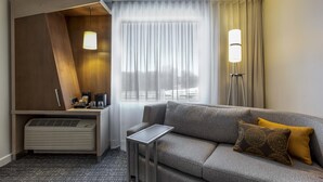 A modern hotel room with a grey sofa, a small table, a desk with a lamp, and a window with sheer curtains. - Courtyard by Marriott Montreal West Island/Baie D'Urfe (Baie-D'Urfe)