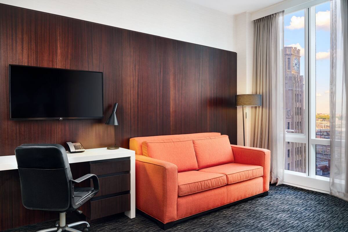 A hotel room with a dark wood panel wall, a flat-screen TV mounted above a desk, an orange sofa, a black office chair, a lamp, and a large window with a city view.