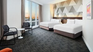 Quarto de hotel com duas camas, uma escrivaninha e uma cadeira. Há uma janela grande que oferece vista da cidade.