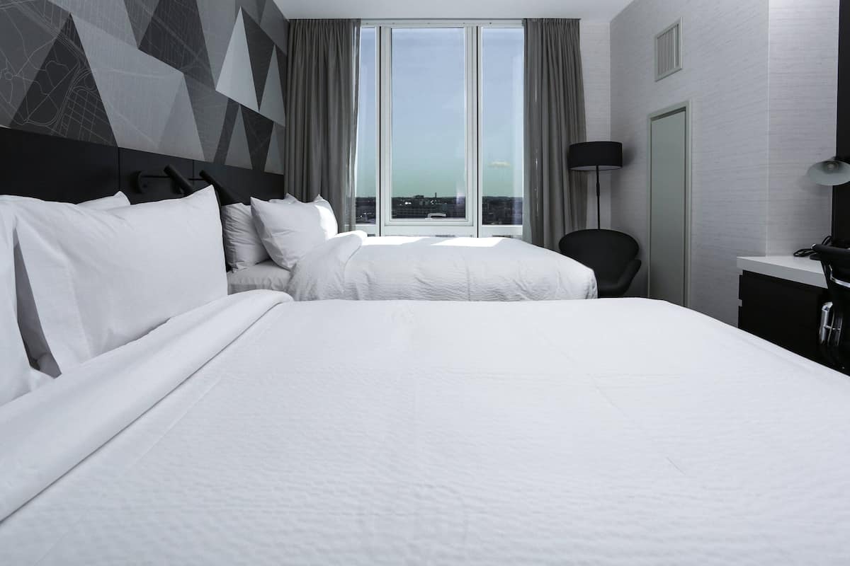A modern hotel room with a large bed, a chair, a desk, and a view of the city through large windows.