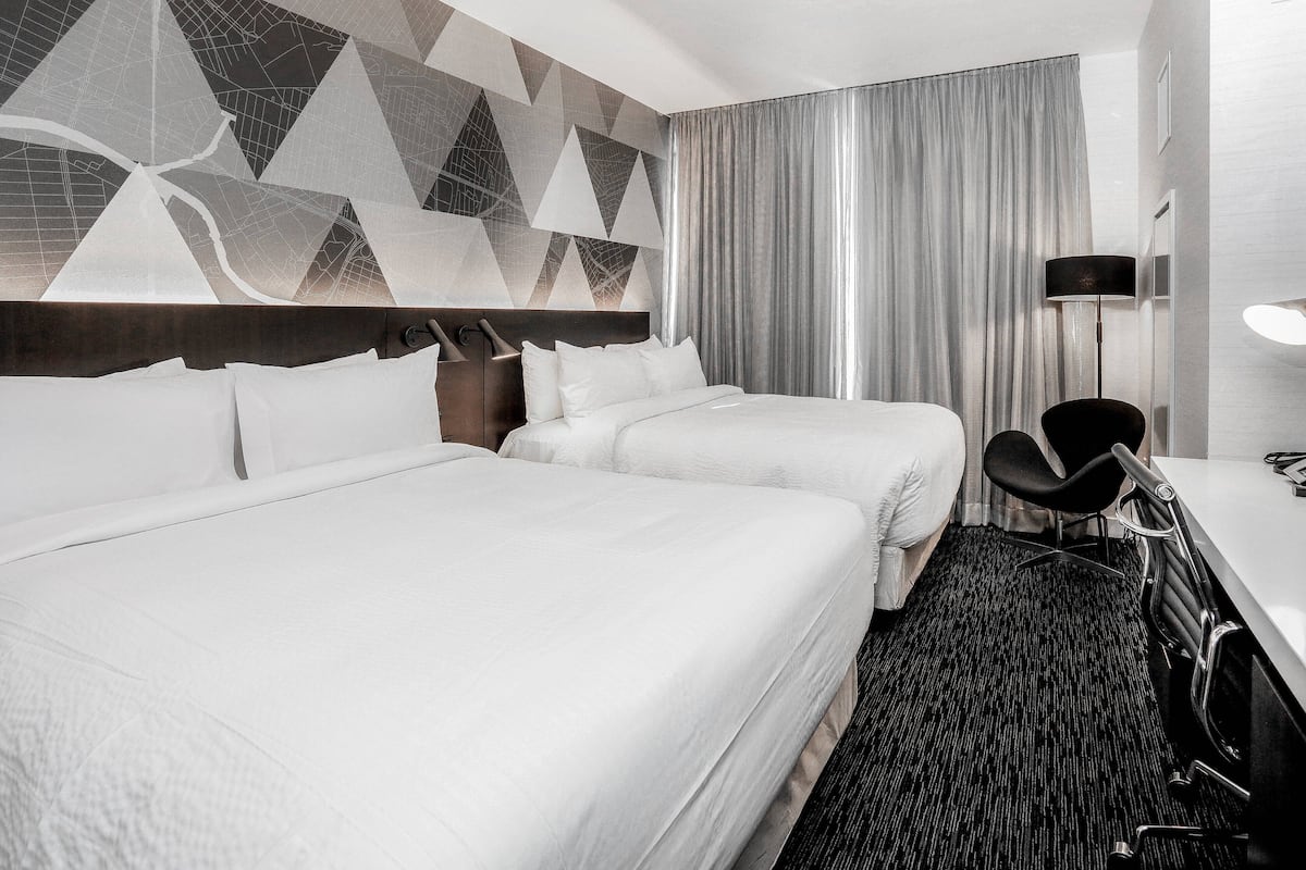 A hotel room with two beds, a desk with a chair, a lamp, and a large window with curtains.