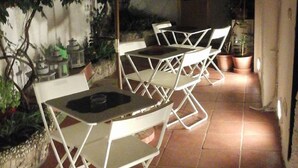Un patio al aire libre con sillas y mesas de metal blanco, un paraguas grande y plantas en macetas.