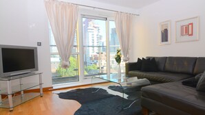 Ein modernes Wohnzimmer mit einem Flachbildfernseher auf einem Glastisch, einem Ledersofa, einem Glastisch und einem schwarz-weißen Teppich. Ein großes Fenster mit Vorhängen rundet das Bild ab.