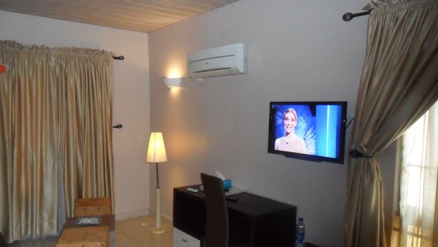 A hotel room with a flat-screen TV mounted on the wall, a desk with a chair, a small ottoman, a lamp, and an air conditioning unit. - Swisscottage suites (Lagos)