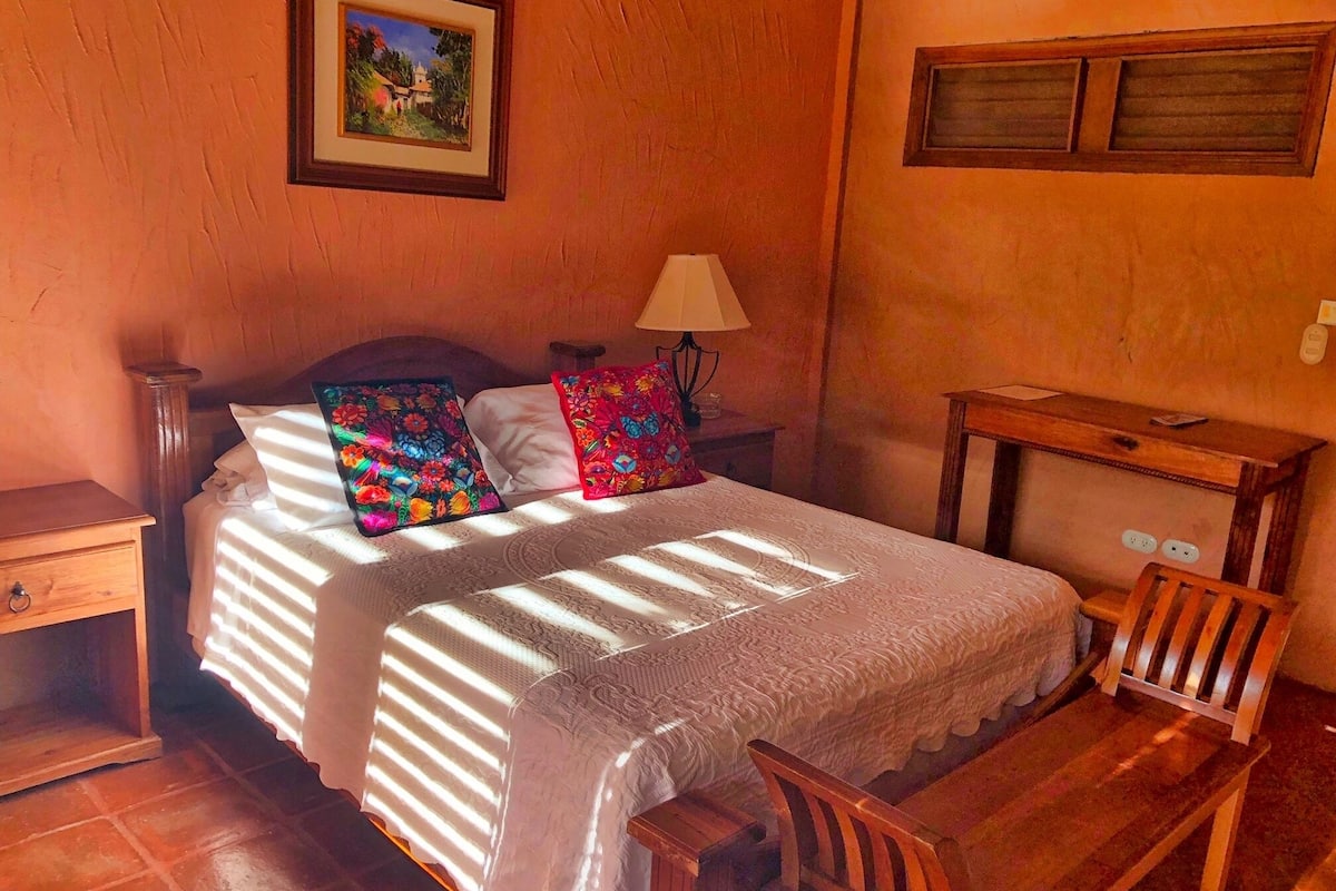 A bedroom with a bed, pillows, a nightstand, a lamp, a wooden chair, a desk, and a window with blinds.
