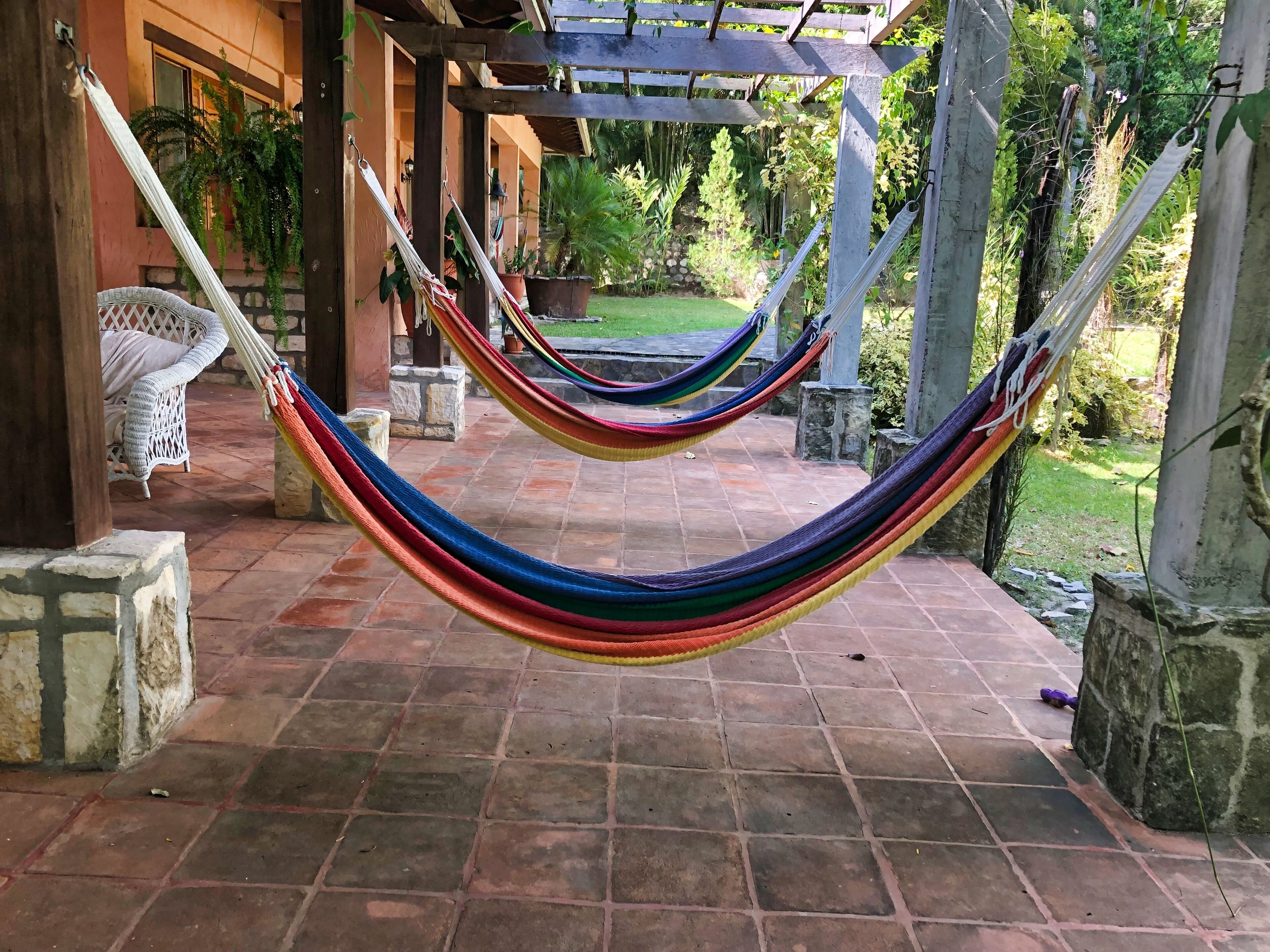 Una hamaca bajo una pérgola de madera, una silla de mimbre y un sendero de piedra que lleva a un jardín.