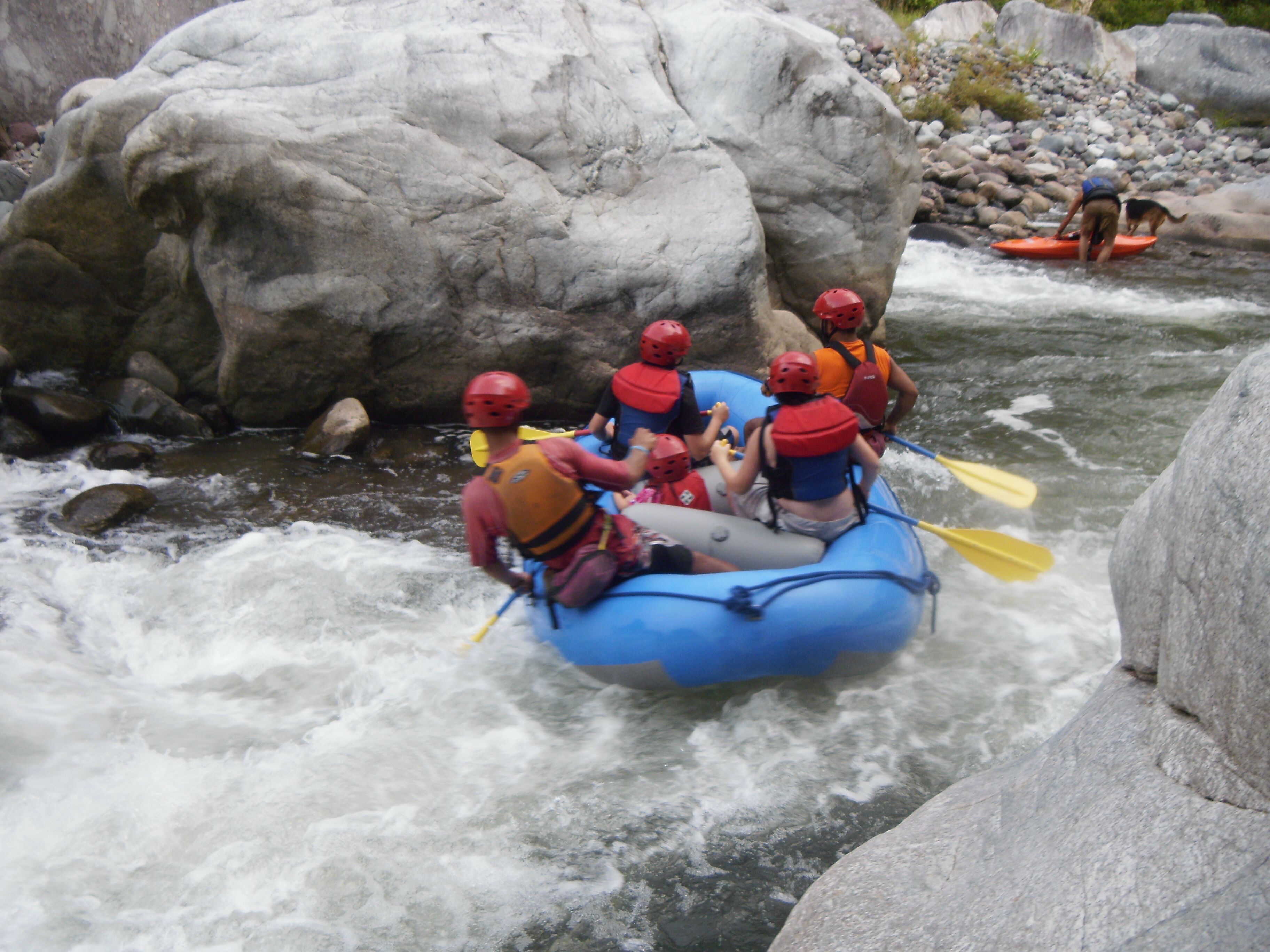 Un groupe de personnes à bord d’un radeau bleu navigue sur une rivière aux eaux tumultueuses, entourée de gros rochers.