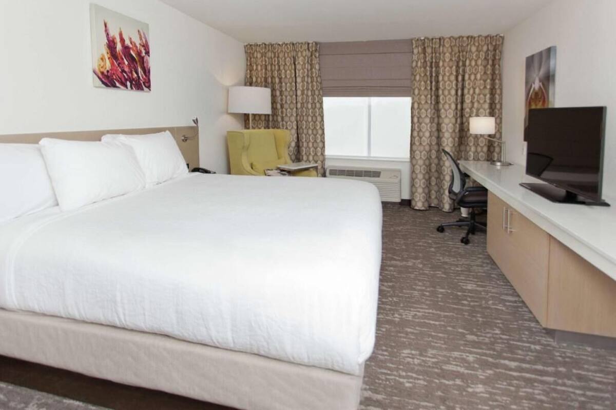 A hotel room with a large bed, a desk with a computer, a chair, a TV, and a window with curtains.