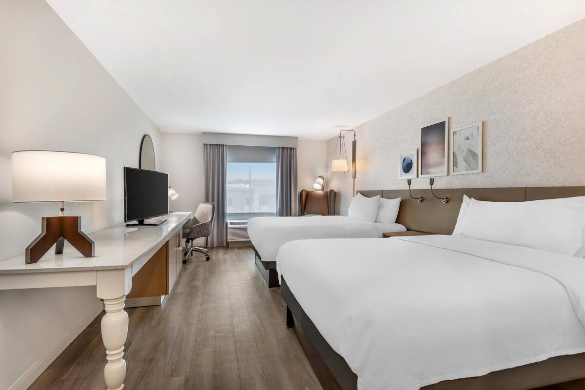 A hotel room with two beds, a desk with a TV, a chair, and a view of the city.