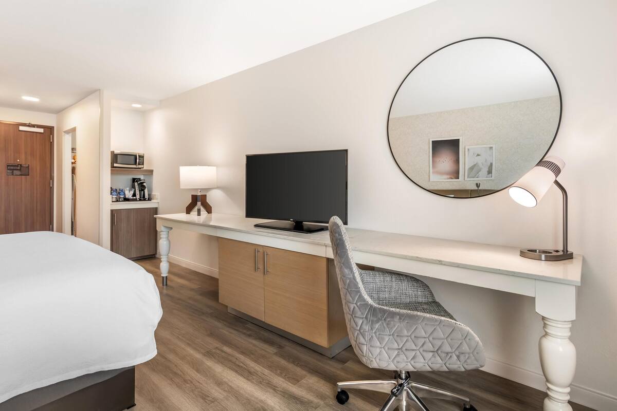 A hotel room with a bed, a desk with a computer, a chair, a round mirror, a lamp, and a microwave.