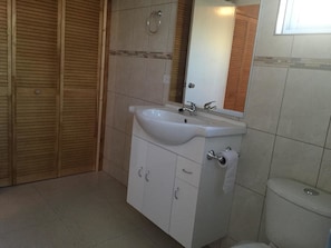 Un baño con lavamanos blanco, espejo y retrete. Hay armarios de madera y un perchero para toallas.