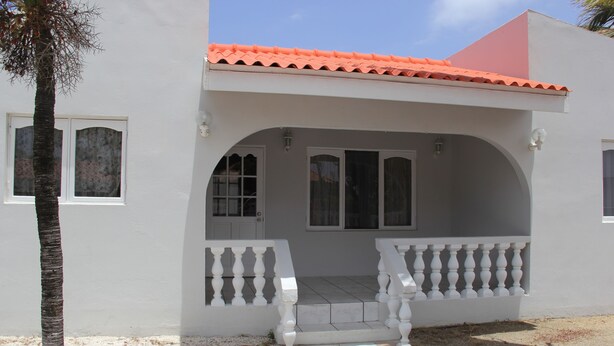 A white building with a red roof, a covered porch with white balustrades, and a small tree in front. - Fitz Aruba 2 Bedroom Home (Noord)