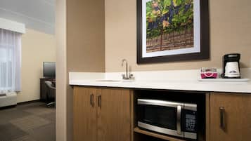 A hotel room with a kitchenette including a microwave, a sink, and a coffee maker. There is a framed picture of a vineyard on the wall.