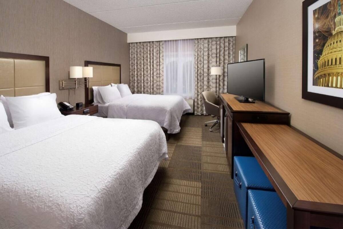 Une chambre d’hôtel avec deux lits, un bureau, une chaise, un téléviseur et un tableau représentant le Capitole.