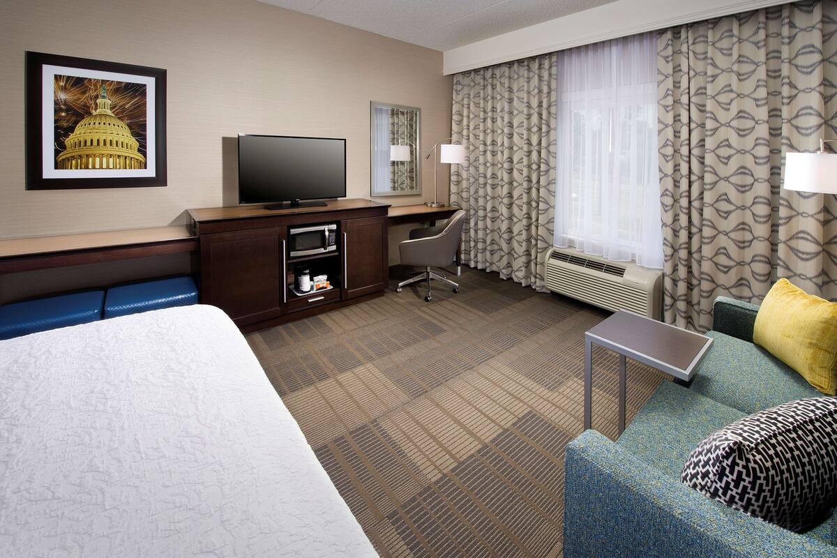 Une chambre d’hôtel avec un grand lit, un bureau avec une télévision, une chaise, une petite table et un canapé avec des coussins.