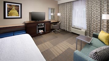 A hotel room with a large bed, a desk with a television, a chair, a small table, and a couch with pillows.