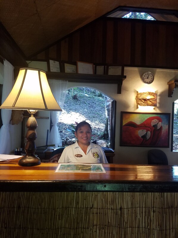 A reception desk with a lamp, a clock, and a painting of parrots. - Table Rock Jungle Lodge (San Ignacio)