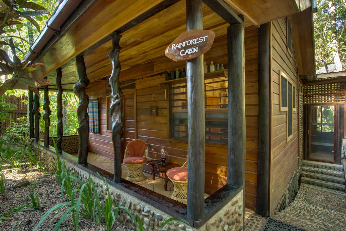 Una cabaña de madera con un porche, identificada como 'Rainforest Cabin', con muebles de mimbre y un sendero de piedra.