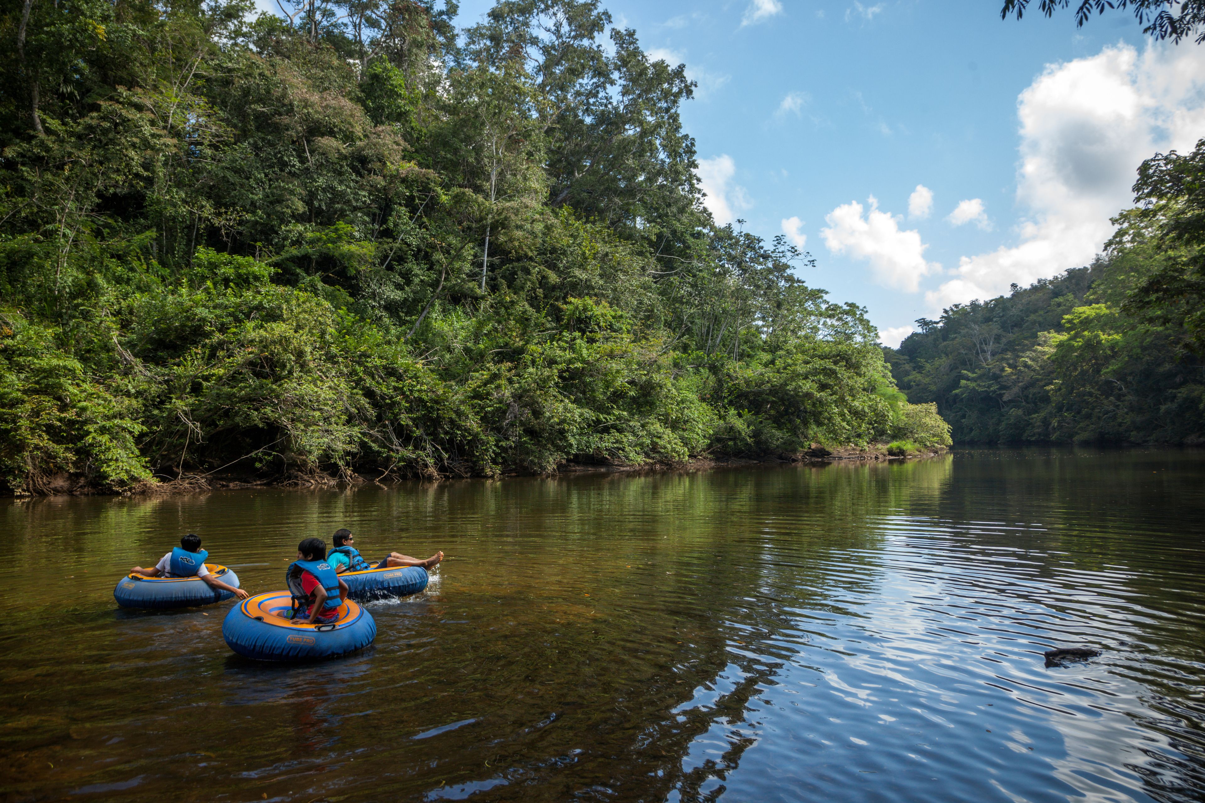 Trois personnes, à l’intérieur de bouées gonflables bleues, flottent sur une rivière paisible, entourée d’une forêt dense.