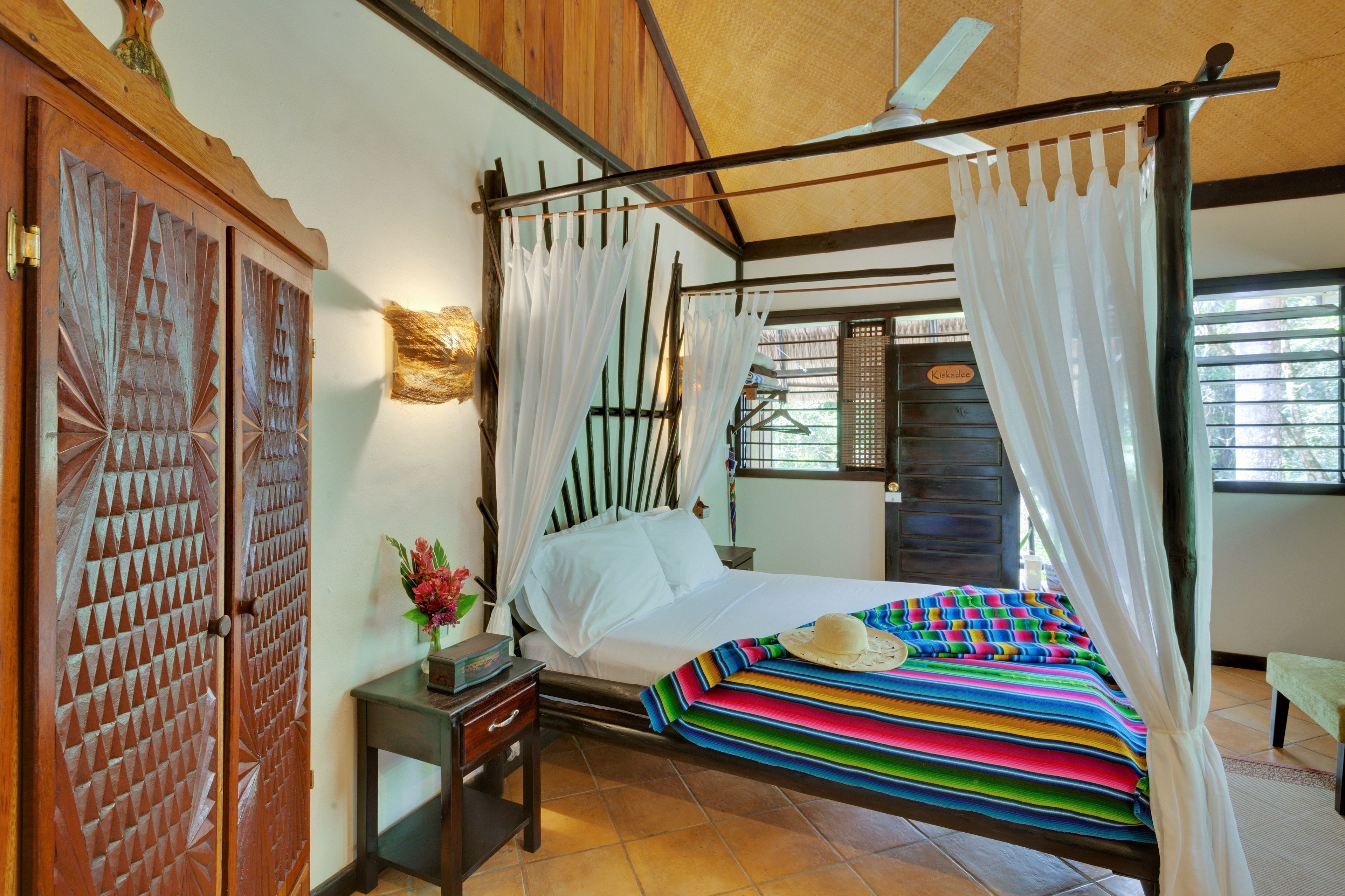 A four-poster bed with white curtains, a colorful striped blanket, and a small table with a vase and book.