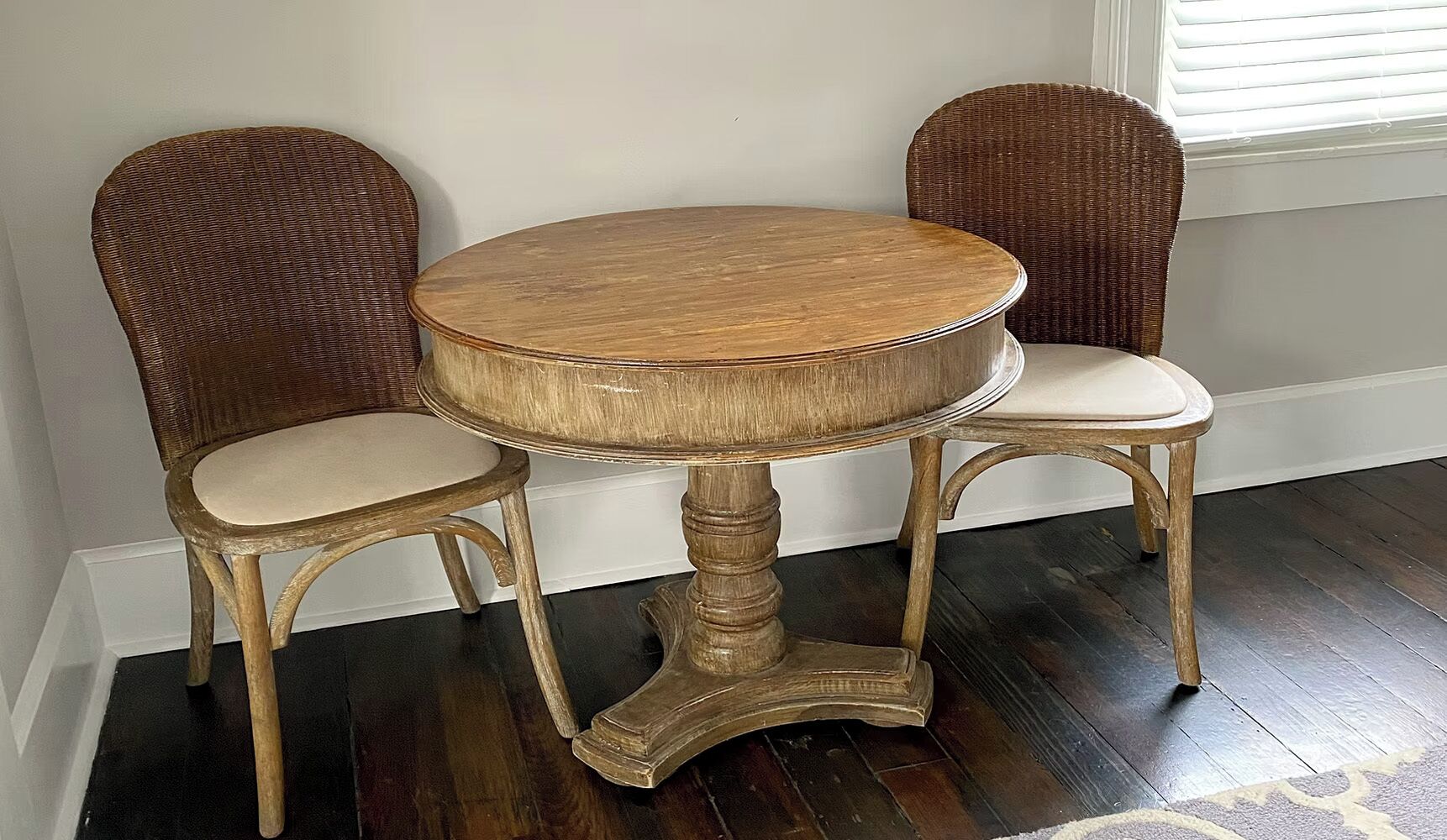 A round wooden table with a pedestal base, flanked by two wicker chairs with cushioned seats.