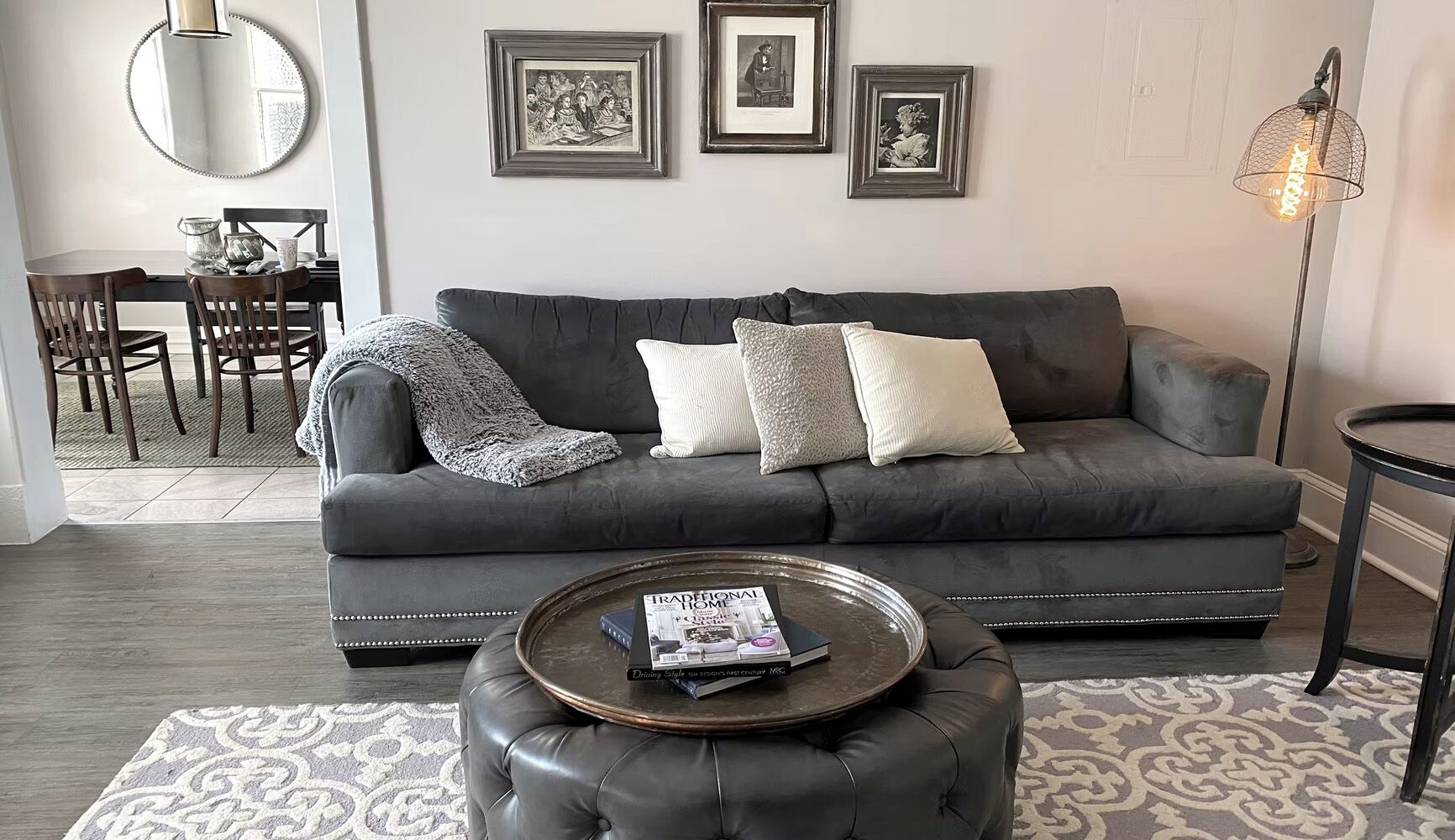 A living room with a dark grey sofa, a round coffee table with a magazine, a patterned rug, and framed pictures on the wall.