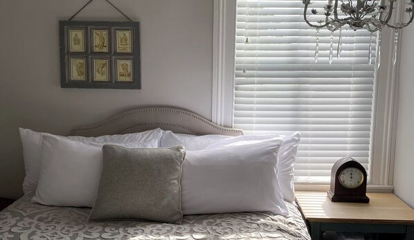 A bed with white and gray bedding, a wall-mounted picture frame, a chandelier, and a bedside table with a clock.