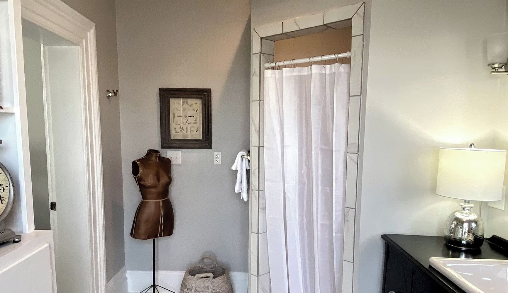 Une salle de bain avec un rideau de douche, un meuble-lavabo avec un miroir, une lampe et un mannequin.