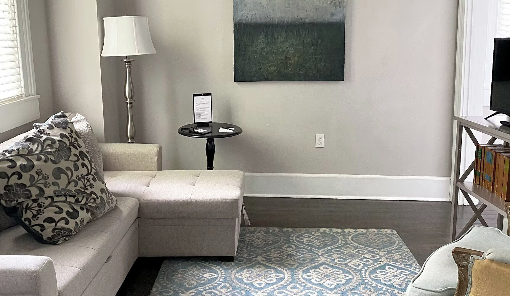 A living room with a sofa, a small table, a lamp, a painting, and a bookshelf.