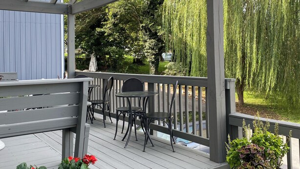Terraza de madera con mobiliario exterior, pérgola y vista a árboles y un coche.