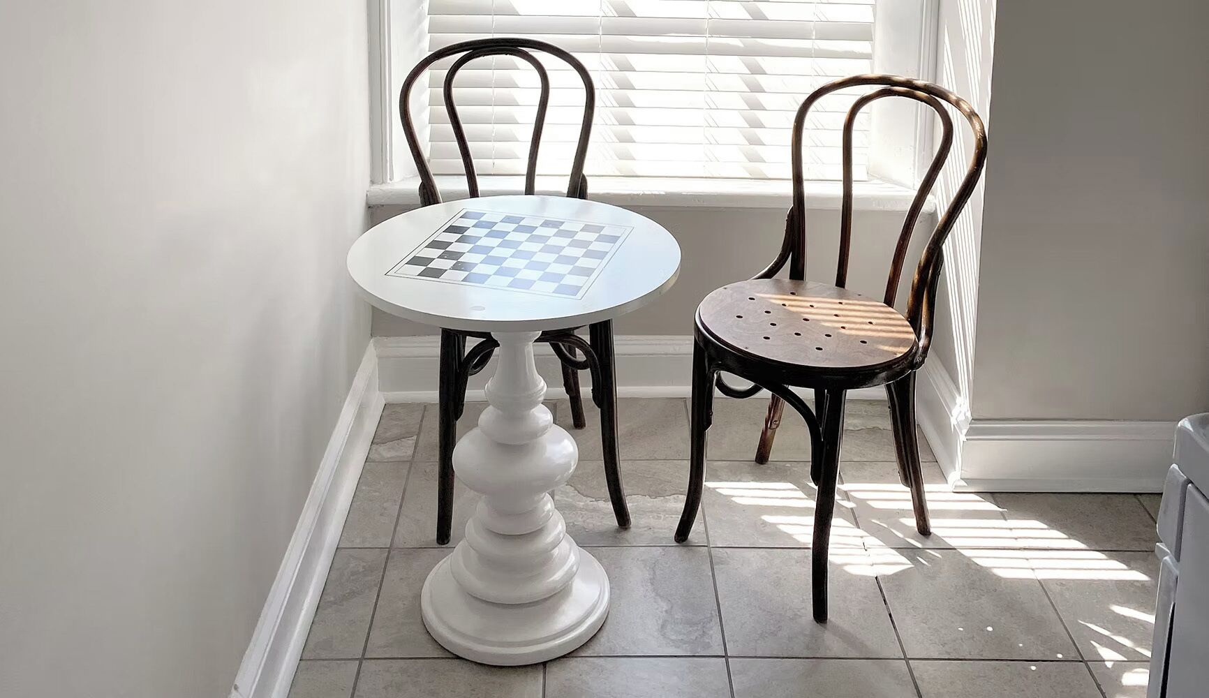 A round table with a chessboard, two chairs, and a window with blinds.
