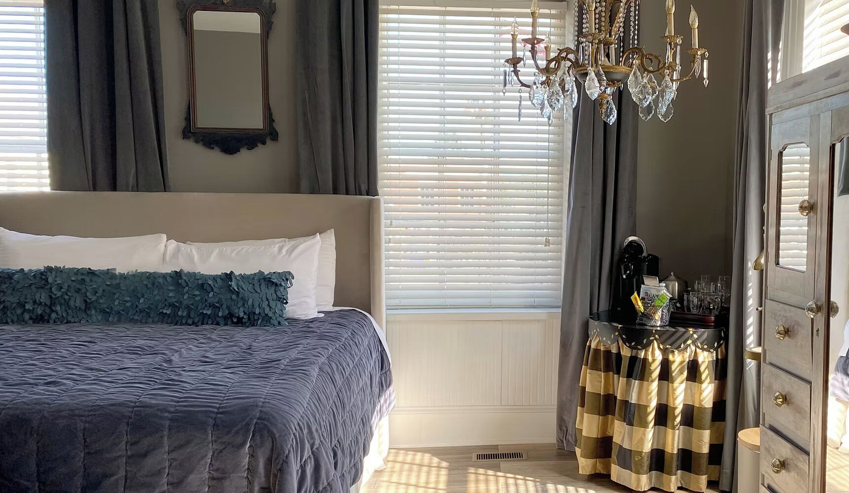 A bedroom with a bed, a dresser, a mirror, a chandelier, and a window with blinds.