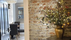Una habitación con pared de ladrillo, zona de comedor con mesa y sillas, y un florero con flores sobre una mesa.