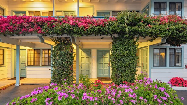 Um edifício de dois andares com varanda coberta, colunas e um canteiro de flores cor-de-rosa.