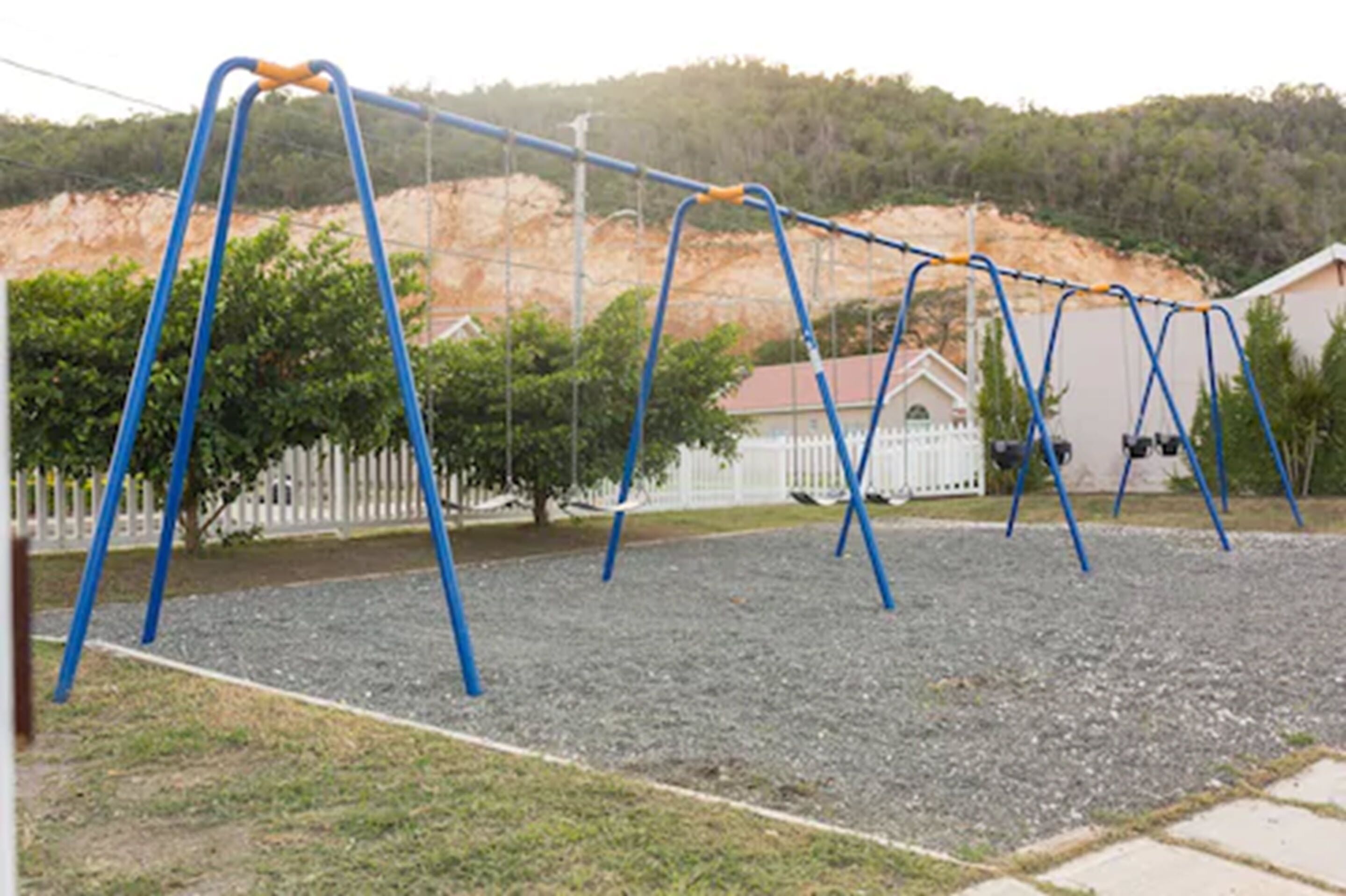 Un terrain de jeux avec des balançoires et une zone délimitée par une clôture, le tout entouré d’une clôture en bois peinte en blanc et d’arbres.