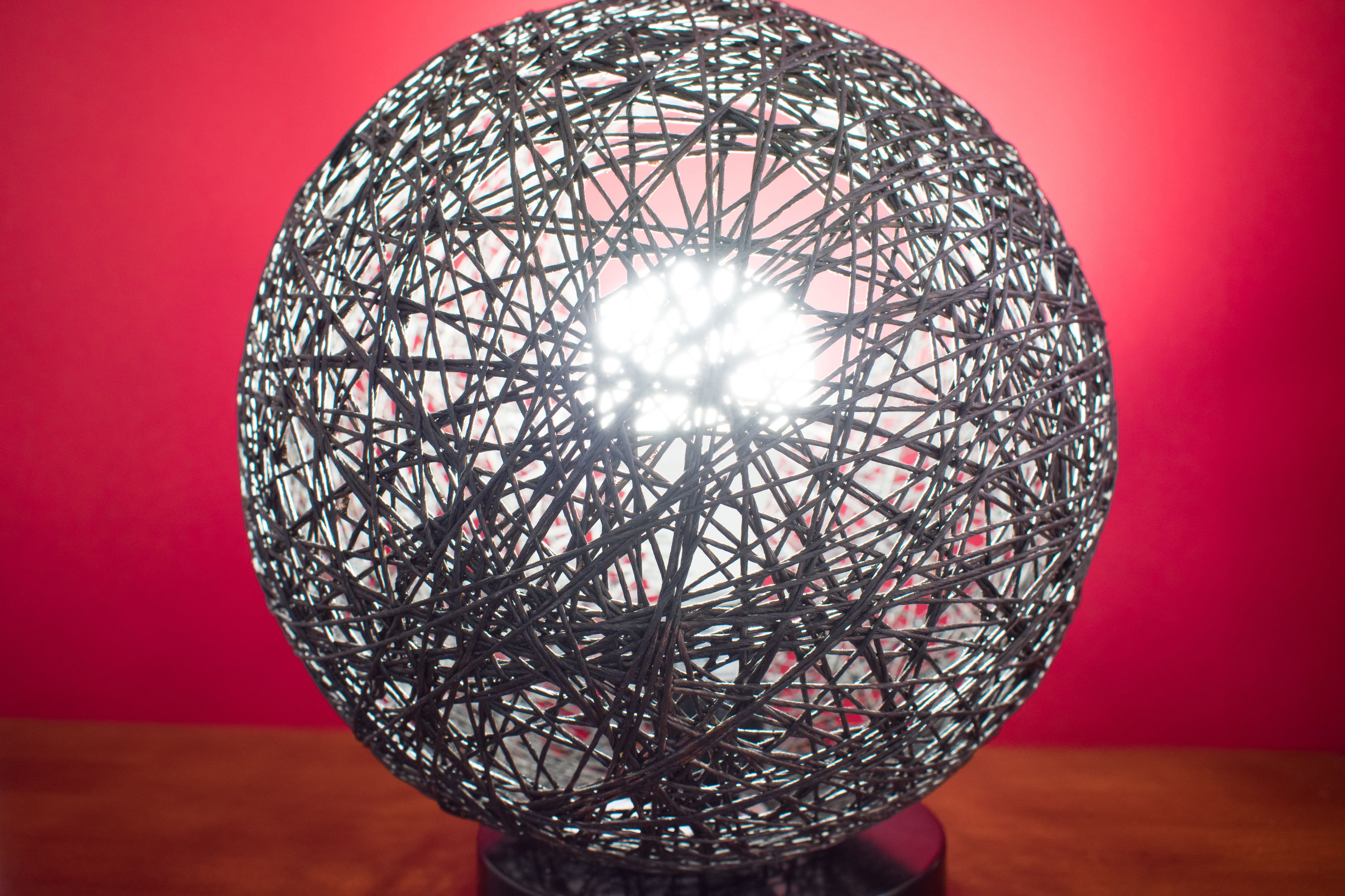 Un objet sphérique doté d’une structure complexe faite de fils métalliques, qui pourrait être un élément décoratif, sur un fond rouge uni.