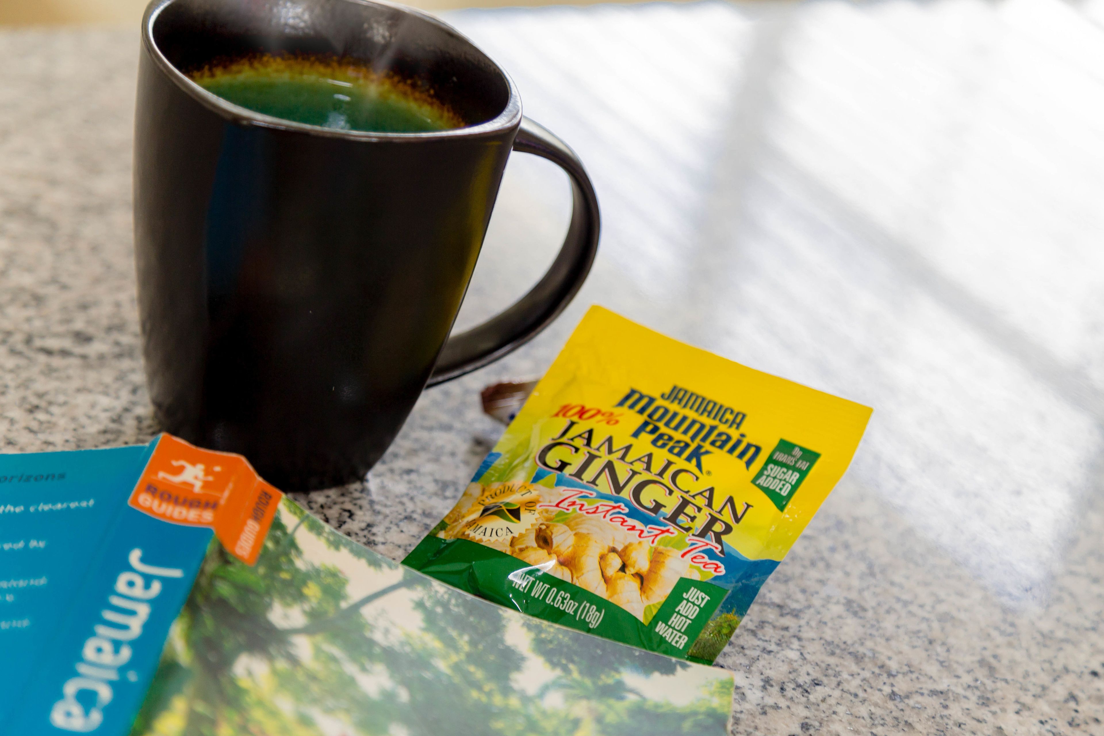 A black mug with a beverage, a packet of Jamaican Mountain Ginger tea, and a travel guide brochure on a countertop.