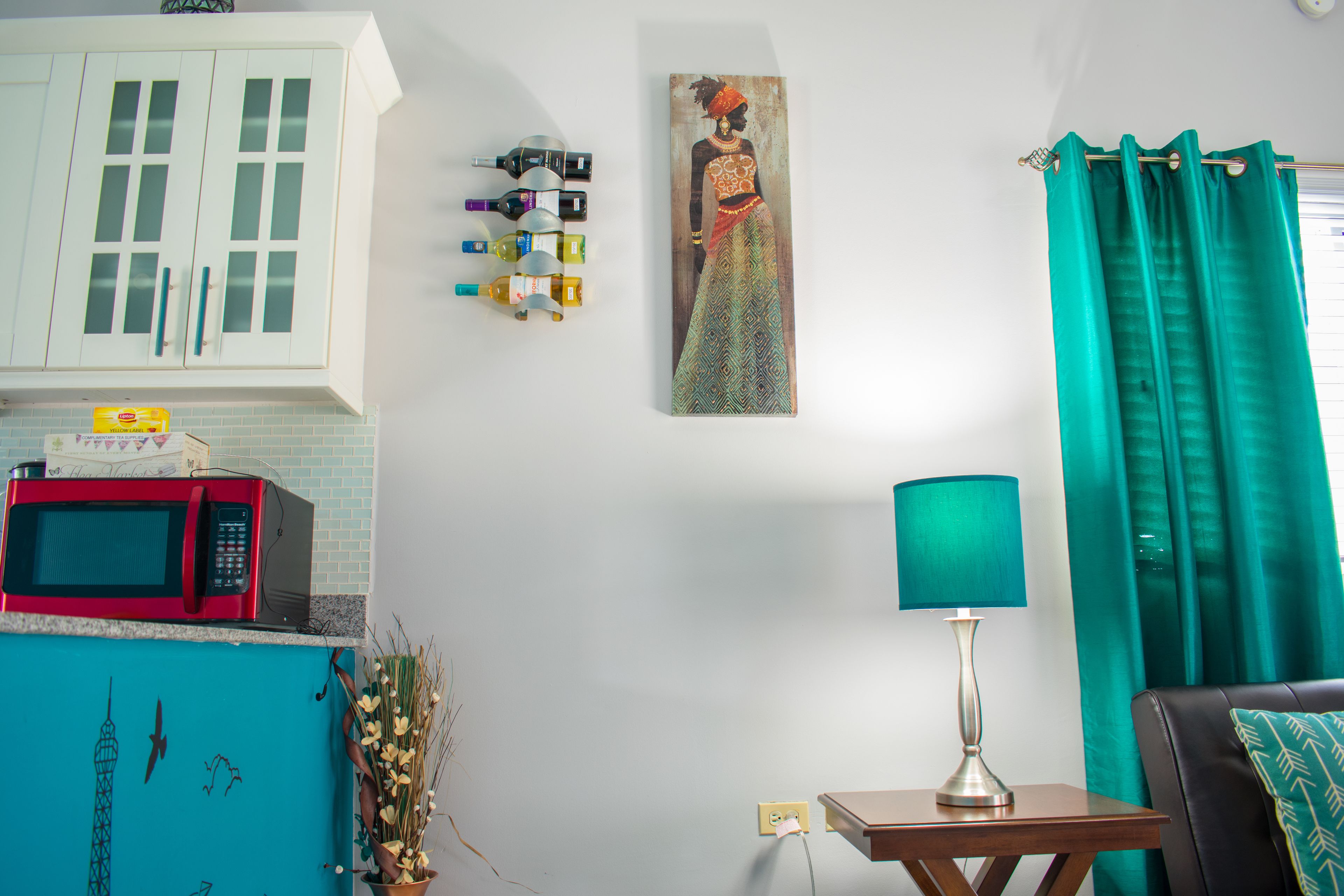 Een keuken met een magnetron, een kast met glazen deuren, een schilderij en een blauwgroen gordijn.